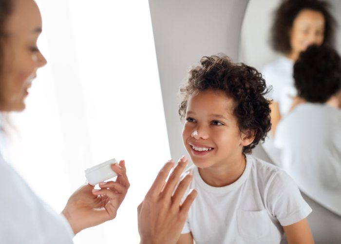 African American young boy is beaming with a joyful smile as his mother gently applies a skincare product to his face. They stand in a well-lit bathroom in front of a mirror